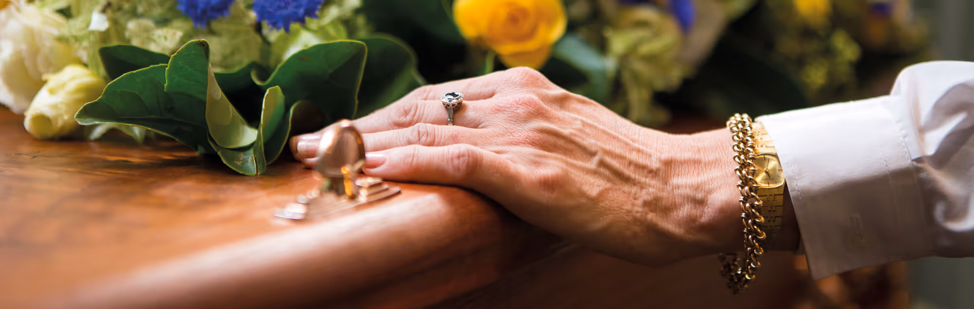 A woman's hand with a ring resting on a wooden casket, adorned with flowers.