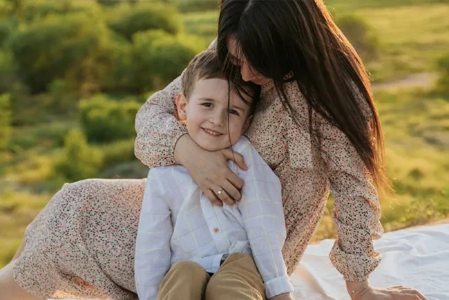 A boy in a white shirt and brown pants smiles while cuddling his mom.