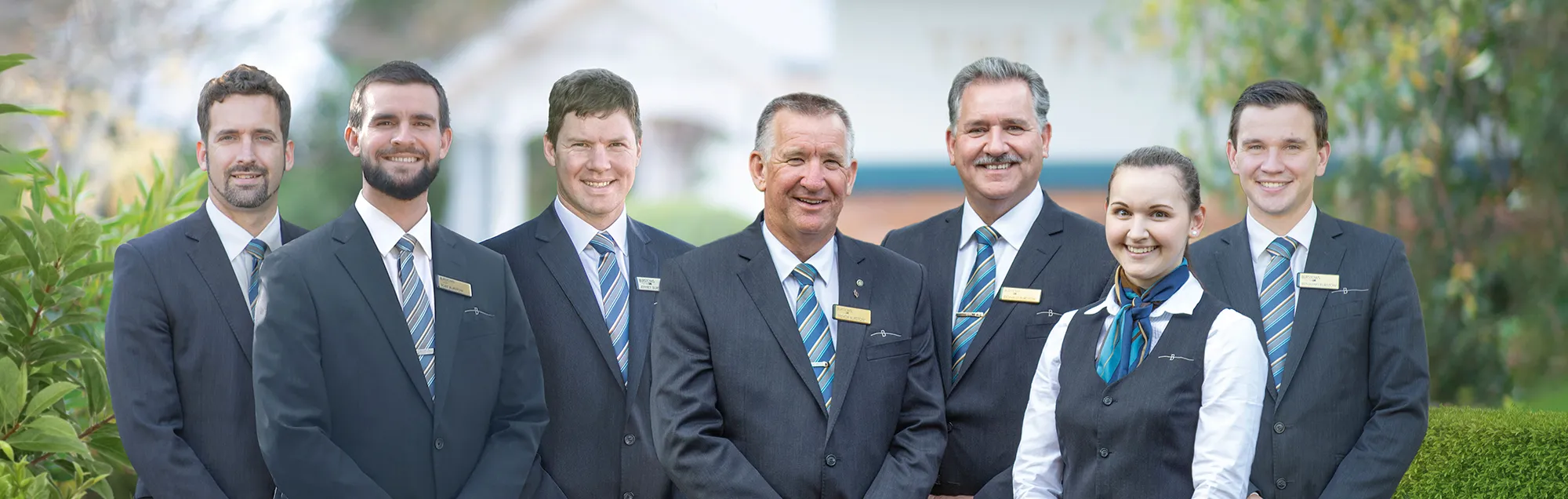A group of six people, five men and one woman, are standing together and smiling at the camera. They are all formally dressed in dark suits, white shirts, and ties, with the woman wearing a vest and scarf. Each person is wearing a name tag on their lapel. The setting appears to be outdoors in front of some buildings and greenery.