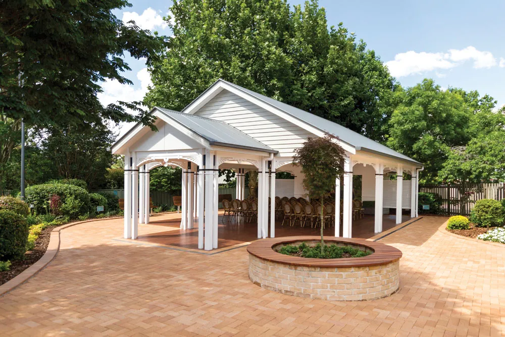 White gazebo with chairs, surrounded by trees and paving.