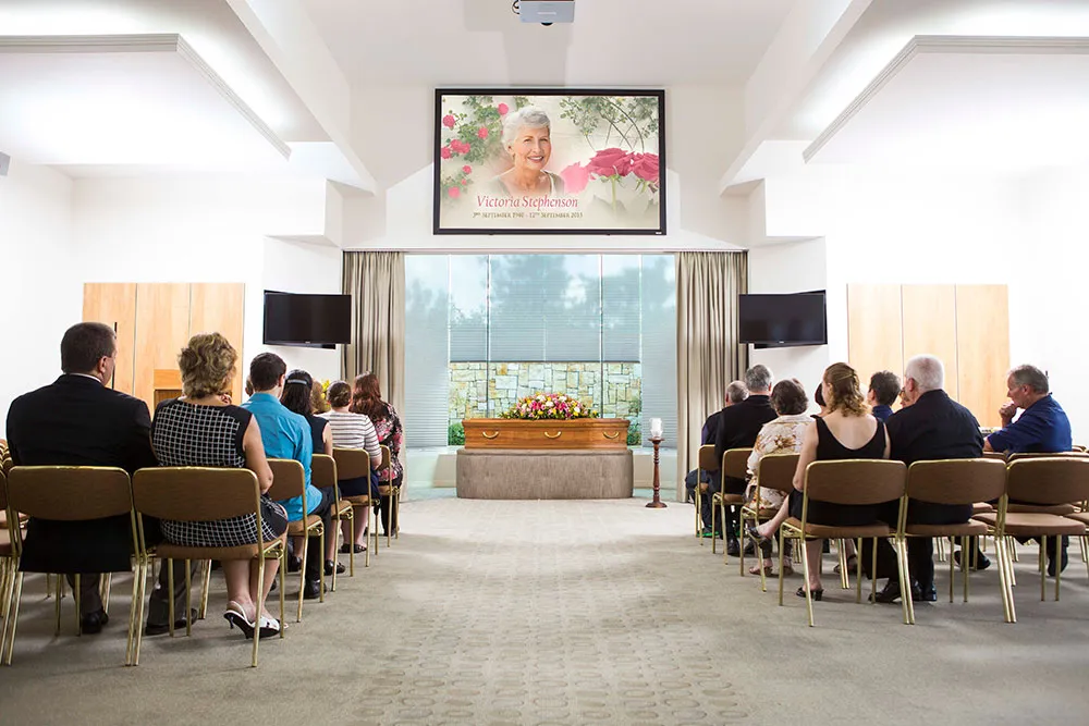 Mourners at a funeral service with a casket and memorial screen.