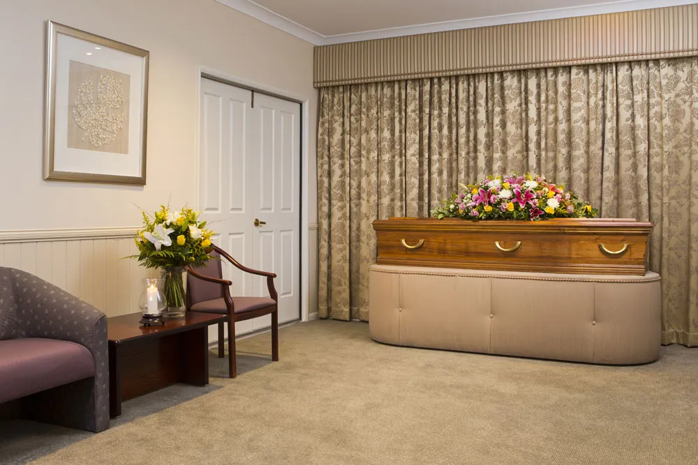 Viewing room with dark furniture, casket, and floral arrangement.